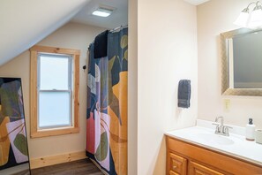 Un baño con ventana, cortina de ducha con estampado de hojas, lavamanos con espejo y una toalla colgada en la pared.