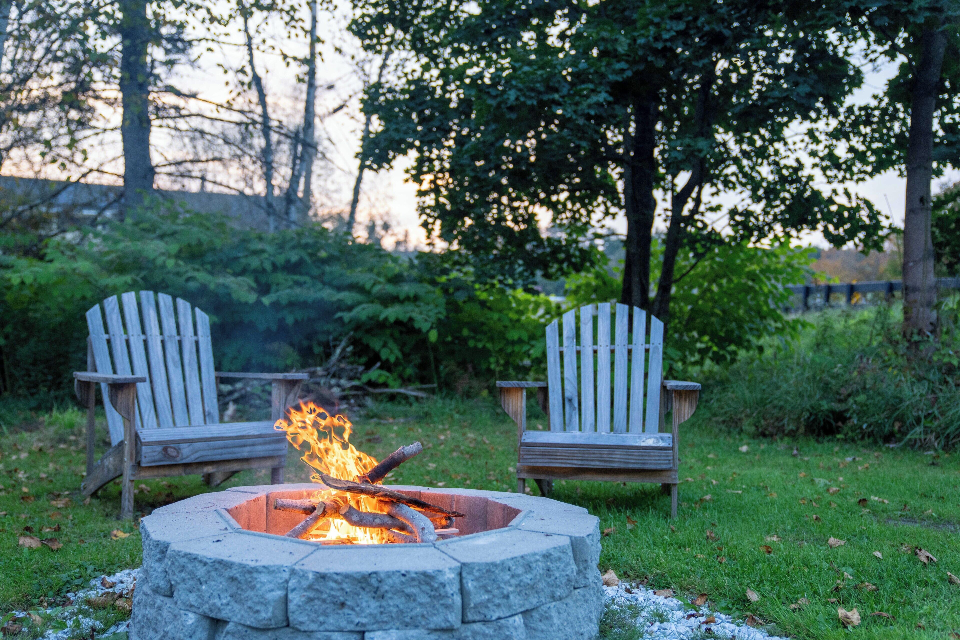 A stone fire pit with burning logs, surrounded by two wooden Adirondack chairs in a grassy outdoor area with trees in the background.