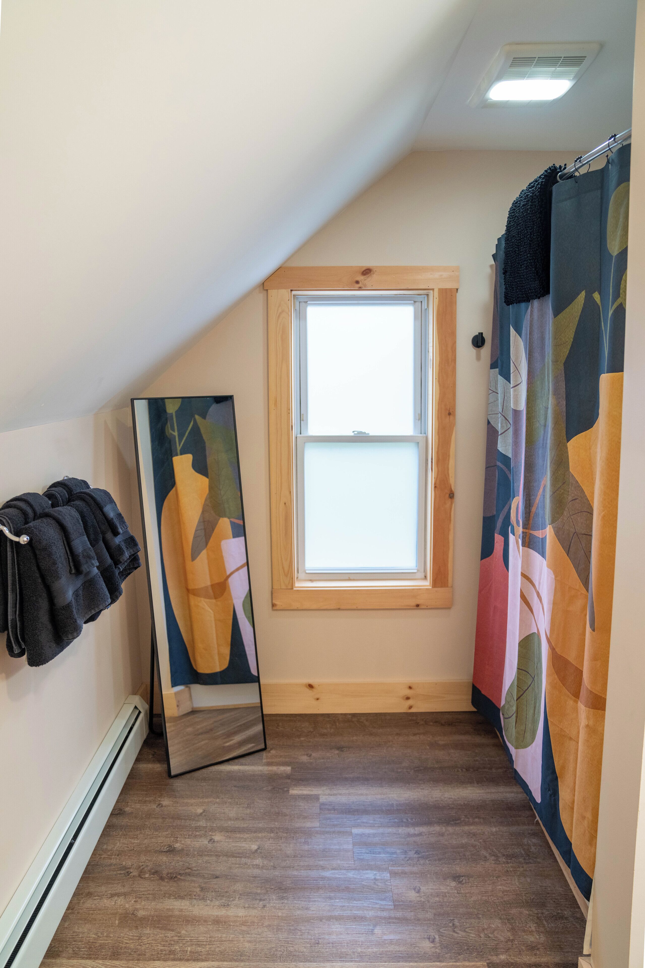 A small, narrow room with a sloped ceiling, a window, a shower curtain, and a mirror leaning against the wall.