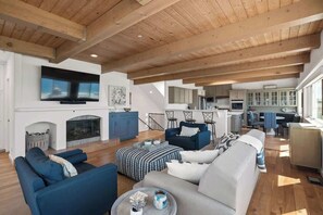 A spacious living room with a fireplace, a television mounted on the wall, and a kitchen area in the background. - Beach House with Spectacular Ocean Views (Manhattan Beach)