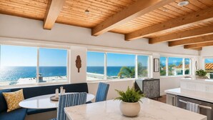 Cozinha moderna com bancada de mármore, área de jantar com mesa redonda e cadeiras azuis, e amplas janelas com vista para o mar.