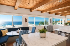 A modern kitchen with a marble countertop, a dining area with a round table and blue chairs, and large windows overlooking the sea. - Beach House with Spectacular Ocean Views (Manhattan Beach)