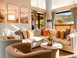 A spacious living room with a sectional sofa, a round coffee table, and a floor lamp. There are two large windows with curtains, and a sign on the wall that reads 'HOT TUB TIME'. - Luxury Tigris House • Hot Tub • King Suites • Near Trails (Sedona)