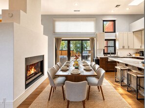 A modern dining room with a fireplace, a large dining table set for six, and a kitchen with white cabinets. - Luxury Tigris House • Hot Tub • King Suites • Near Trails (Sedona)