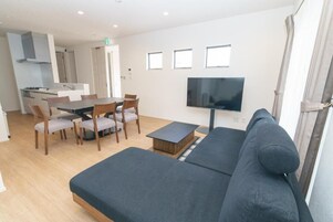 A modern living room with a sofa, a dining table with chairs, a flat-screen TV, and a kitchen area in the background.