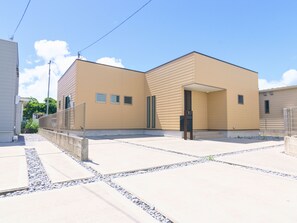 A modern building with a flat roof and a beige exterior. There are three windows on the front facade, and a door is visible on the right side. The building is surrounded by a paved area with small pebbles and concrete slabs.