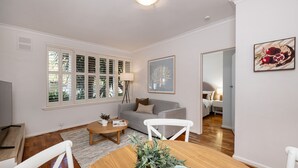Een moderne woonkamer met een bank, een salontafel en een eettafel. Er is een raam met zonwering, een lamp en een ingelijst schilderij aan de muur.