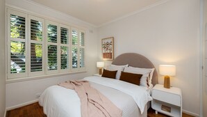 Een slaapkamer met een groot bed, twee nachtkastjes met lampen, een raam met jaloezieën en een schilderij aan de muur.