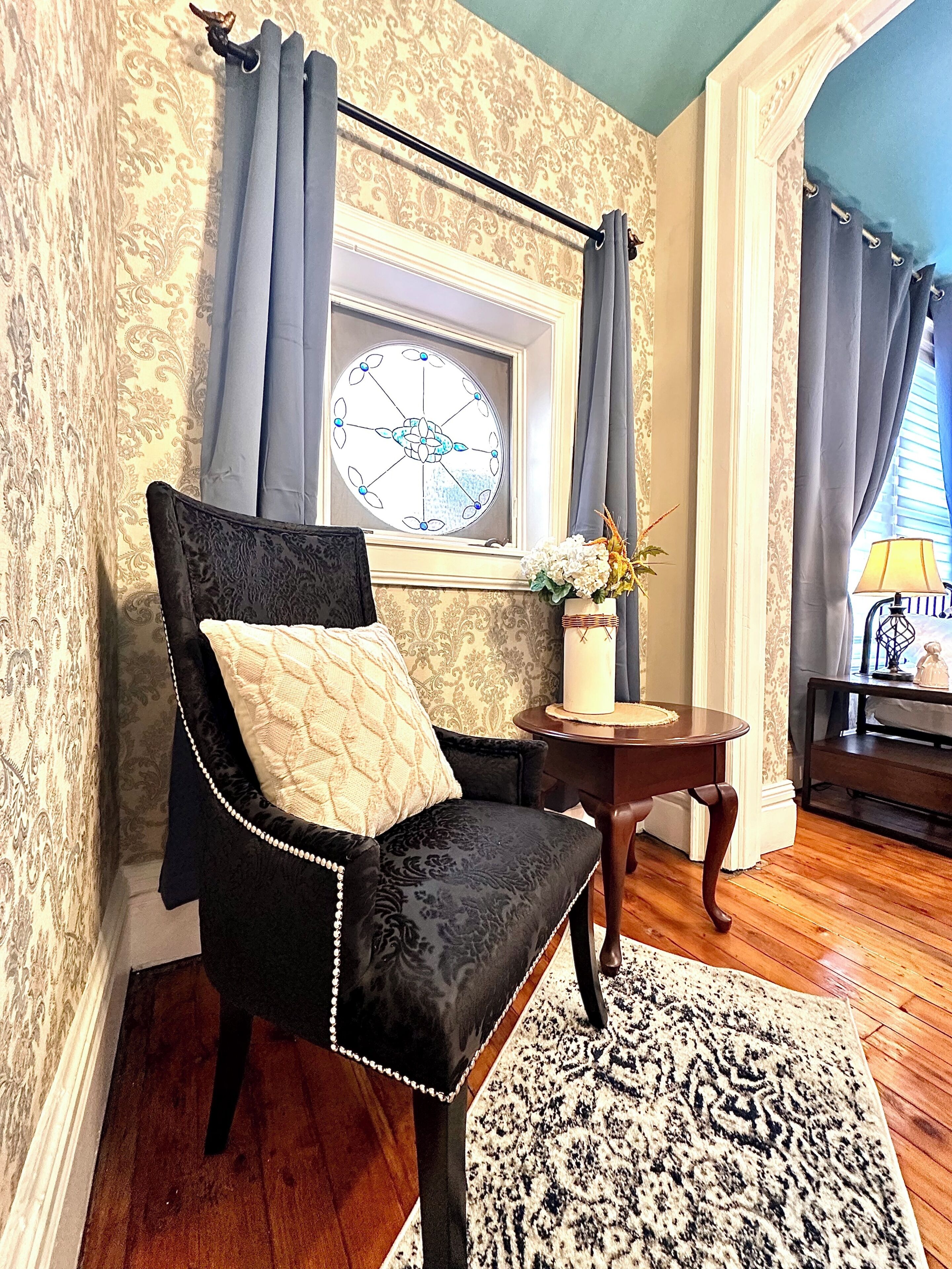 A room with a black upholstered chair, a round wooden table, a patterned rug, and a window with a decorative glass.