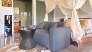 A modern bedroom with a grey sofa, a glass coffee table, and a bed with a canopy. - Rana Suite (Ubud)