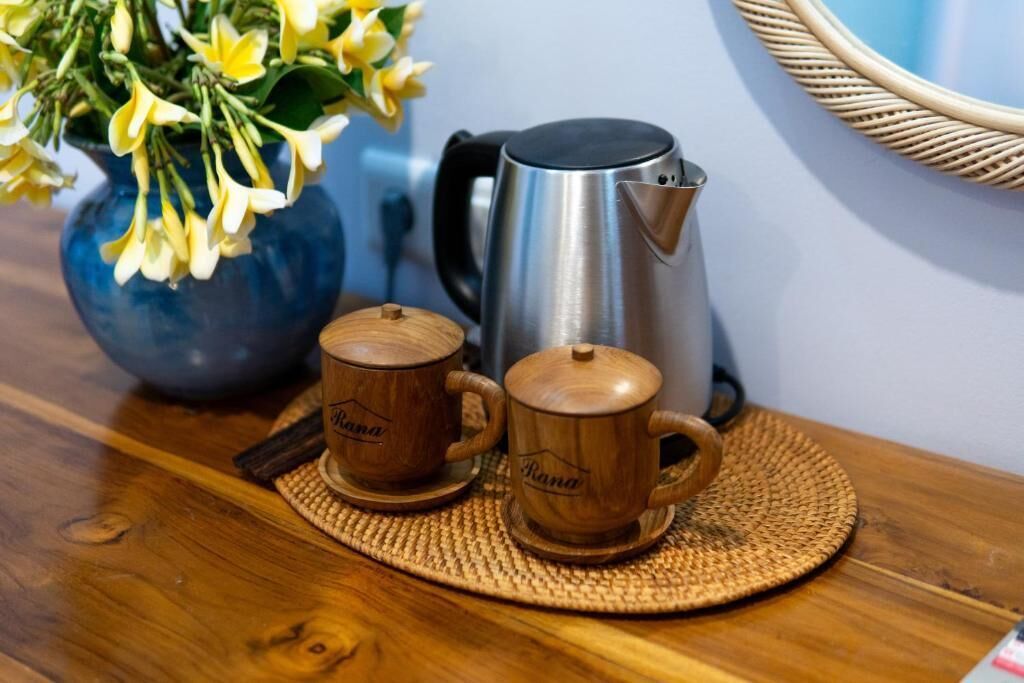 Two wooden cups with lids on a woven placemat, a stainless steel kettle, and a blue vase with yellow flowers on a wooden table.