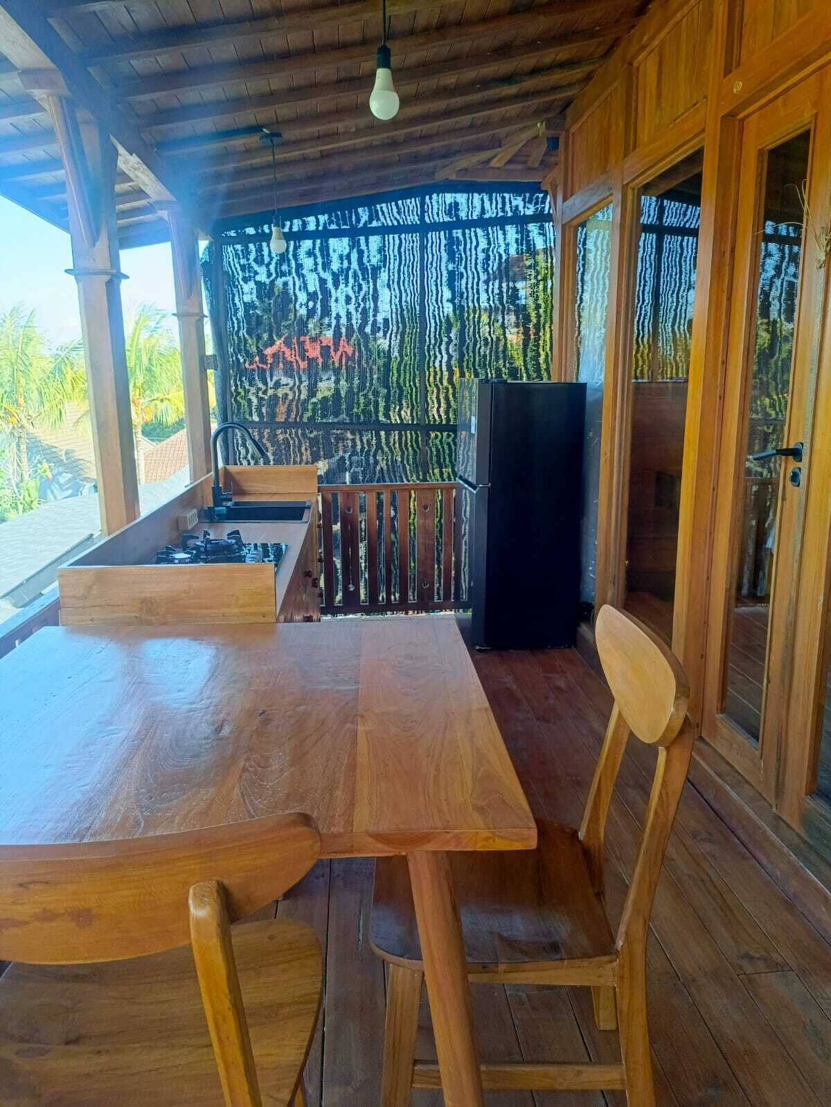 A wooden deck with a table and chairs, a refrigerator, and a view of palm trees.
