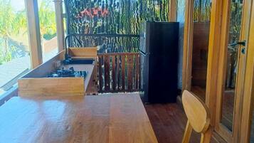 A wooden deck with a table and chairs, a refrigerator, and a view of palm trees.