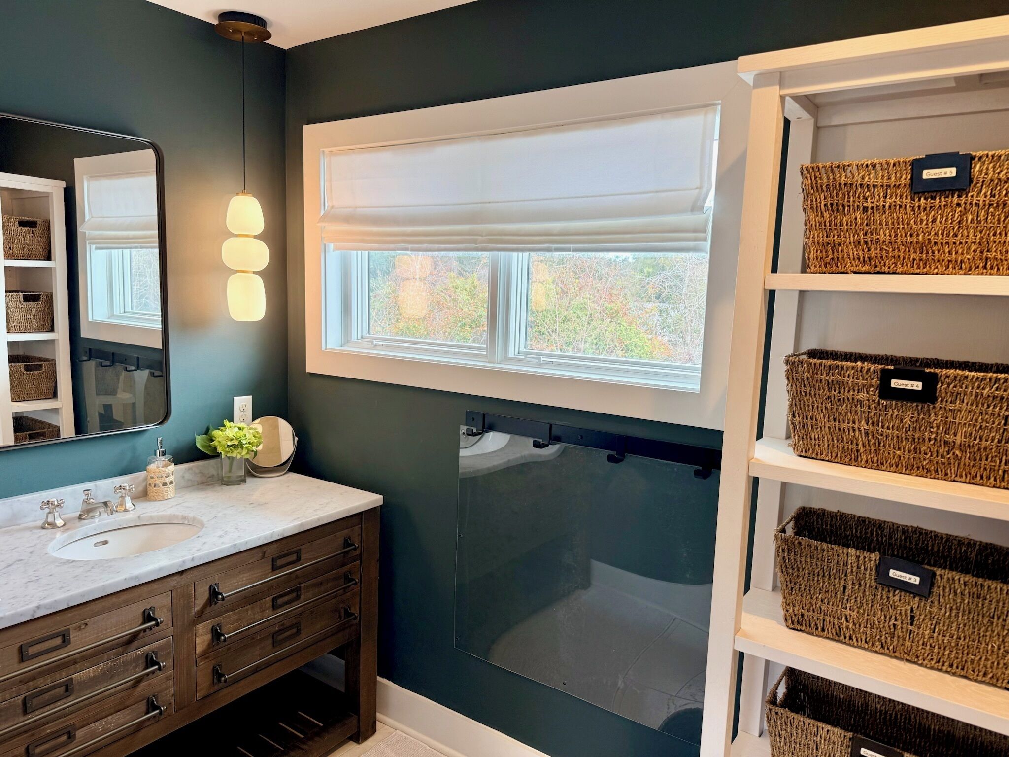 A bathroom with a dark teal wall, a white vanity, a large mirror, and a window above the sink. There are wicker baskets labeled "Guest # 5" and "Guest # 4" on shelves.