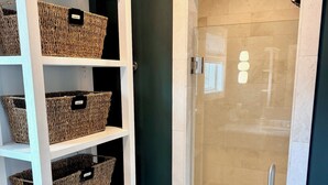 Un baño con un estante blanco que contiene cestas de mimbre, una cabina de ducha de cristal y una pared en color verde azulado oscuro.