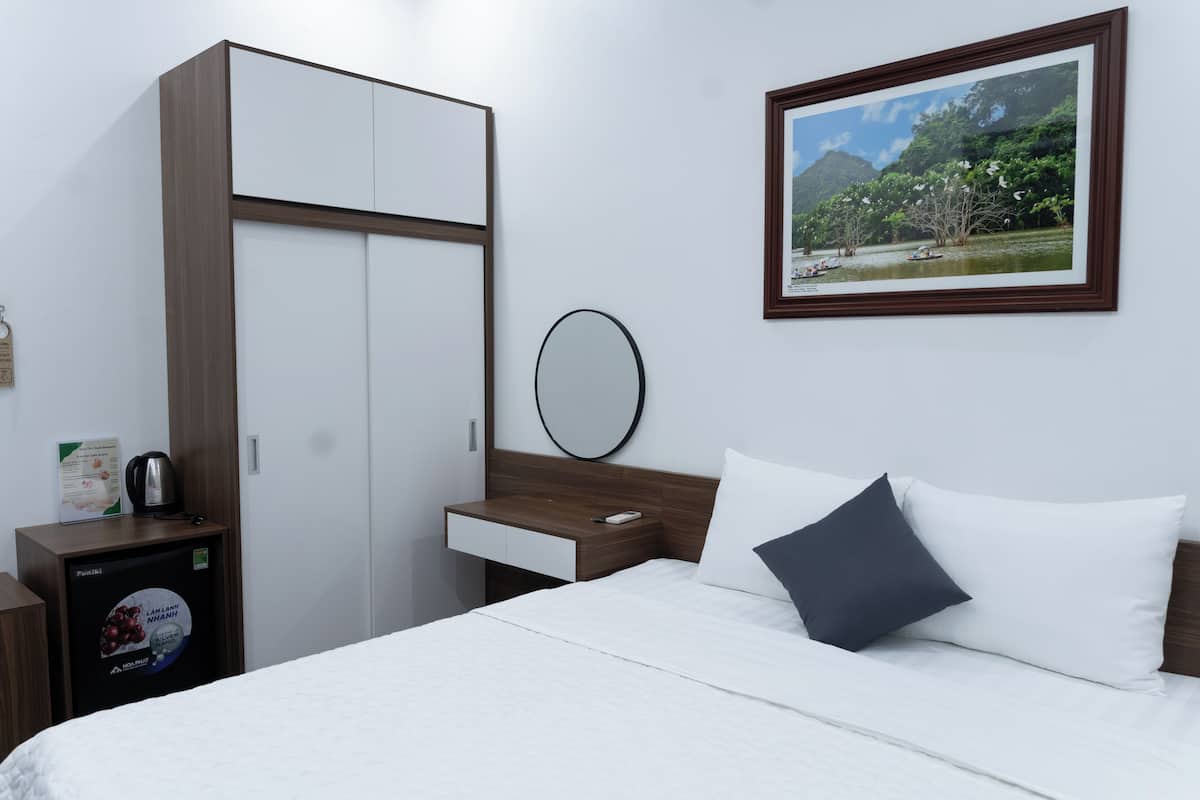 A hotel room with a bed, a wardrobe, a small table with a kettle, a framed picture on the wall, and a bedside table with a remote control.