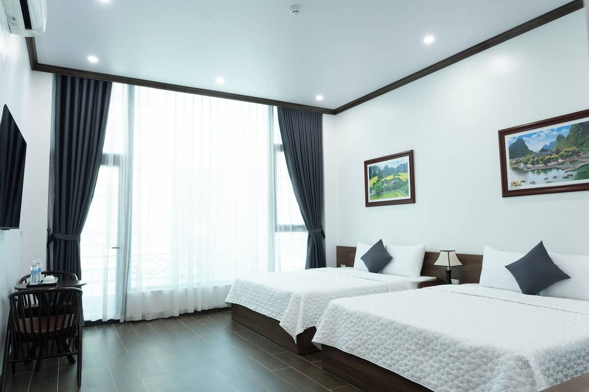 A hotel room with two beds, a desk, and a chair. There are curtains, a painting on the wall, and a window with a view.