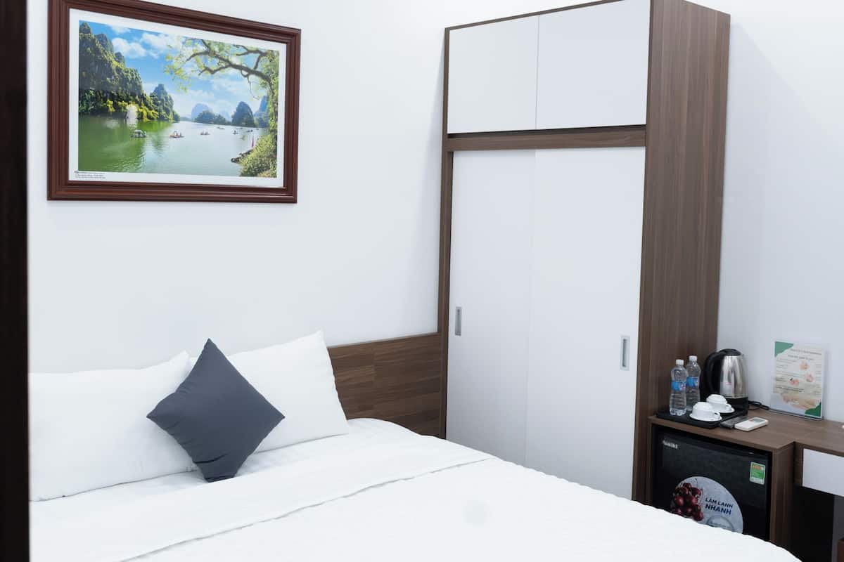 A neatly made bed with a white duvet and a dark pillow, a wooden wardrobe, a small table with a kettle and a water bottle, and a framed picture of a landscape on the wall.