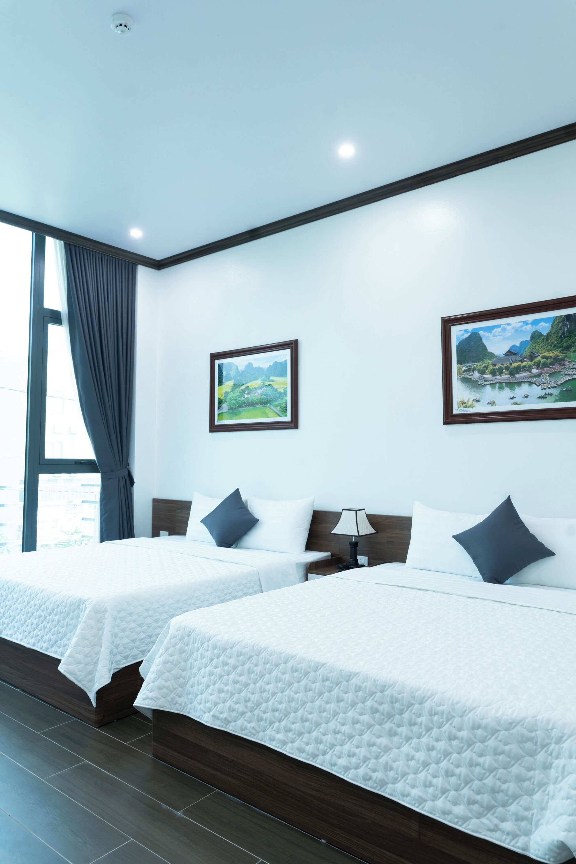 A hotel room with two beds, a large window with curtains, and two framed pictures on the wall.