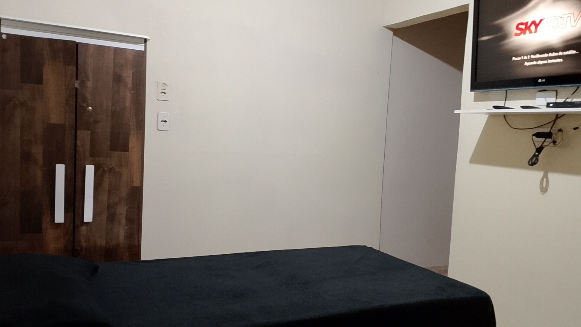 Een kamer met een houten garderobe, een wandgemonteerde flatscreen televisie en een bed met donkere beddengoed.