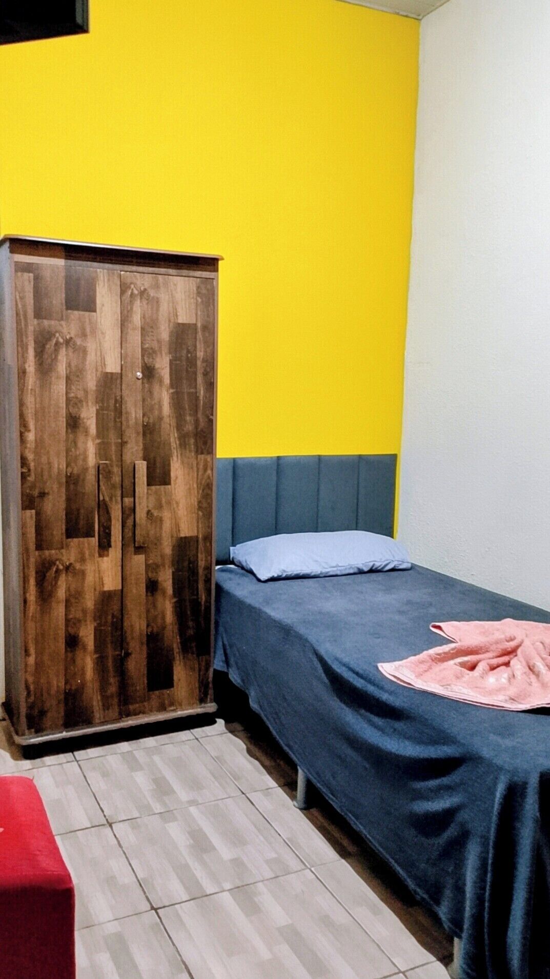 A room with a wooden wardrobe, a bed with a blue headboard, and a red chair.