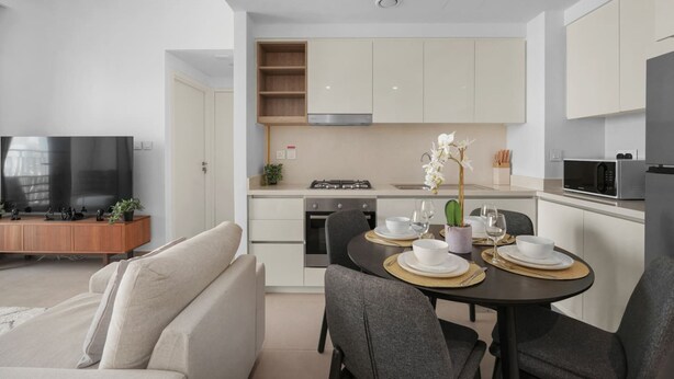 A modern living room with a sofa, a dining table set for two, and a kitchen area with cabinets, a stove, and a microwave. - 1BR Retreat Near Burj Khalifa (Dubai)