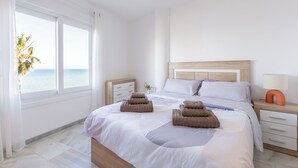 Ett sovrum med en säng, sängbord, ett litet bord, en lampa och utsikt över havet genom stora fönster.