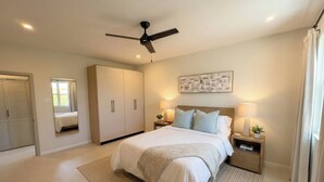 Ein Schlafzimmer mit einem Bett, Nachttischen, einem Deckenventilator, einem großen Fenster mit Vorhängen und einem an der Wand hängenden Kunstwerk.
