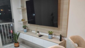 A modern living room with a large flat-screen TV mounted on the wall, a white console table with decorative items, two beige armchairs, and a potted plant on the floor. - New apartment well located in a 4 floor brand new with all the comforts. (Barranquilla)