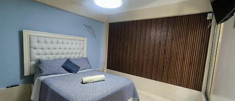 Une chambre à coucher avec une tête de lit capitonnée, une couvre-lit à motifs, un ventilateur de plafond et un mur recouvert de panneaux en bois.