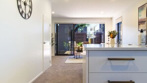 Cocina moderna con una isla grande, un reloj en la pared y un muro de vidrio corredizo que da a un espacio exterior con vistas a edificios.