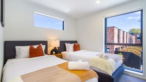Habitación de hotel con dos camas, una mesita de noche con lámpara y ventanas amplias que dan a un edificio de ladrillo.