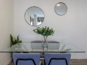 A modern dining area with a glass table, two grey chairs, a vase with green plants, and two round mirrors on the wall.