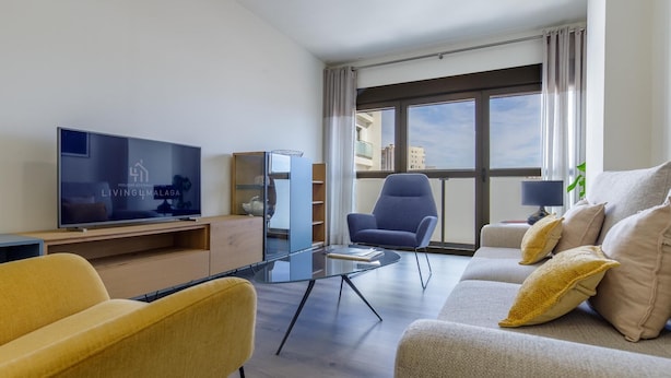 Un salon moderne avec un téléviseur à écran plat, un canapé, un fauteuil jaune, une table basse en verre et une grande fenêtre avec des rideaux.