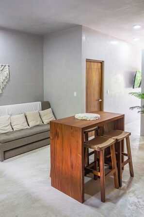 Ein modernes Wohnzimmer mit einem Holztisch, zwei Barhocker, einem grauen Sofa mit Kissen und einer Holztür.