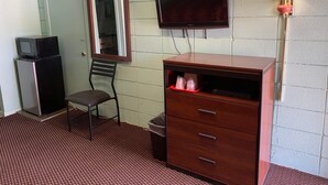 A room with a television, a desk with drawers, a chair, a trash bin, and a lamp.