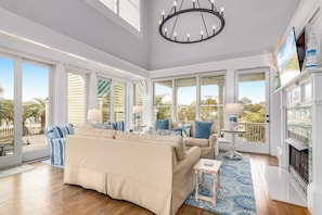 A spacious living room with large windows, a fireplace, and a chandelier. - Seas the Day delivers Atlantic Ocean seascapes (Bald Head Island)