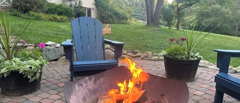 A metal fire pit with flames, surrounded by potted plants and blue chairs on a paved patio.