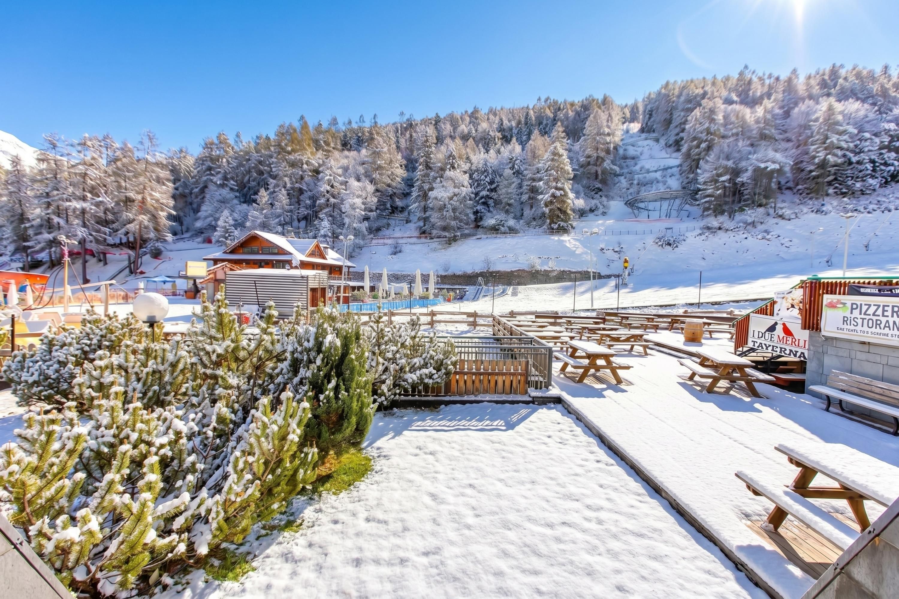 Un espace extérieur enneigé avec des tables et des bancs de pique-nique en bois, un chalet en bois et un panneau indiquant une pizzeria et un restaurant.