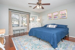 A bedroom with a bed, a ceiling fan, a window with blinds, a dresser, and framed pictures on the wall.