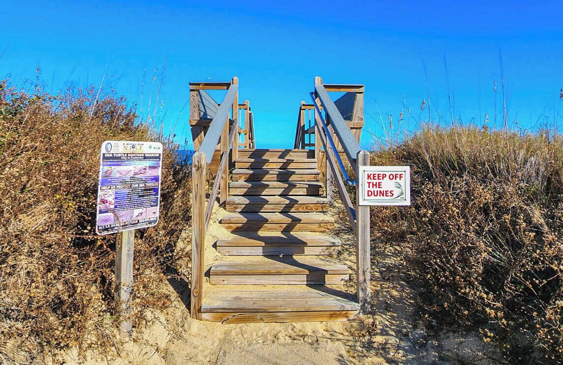 A wooden staircase leading to beach access, with signs indicating a sea turtle nesting season and a 'Keep Off The Dunes' warning.