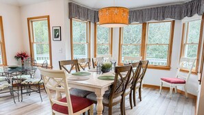 Uma área de refeições com mesa e cadeiras de madeira, janelas amplas e um lustre.