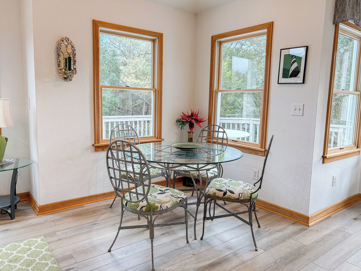 Un coin repas avec une table ronde en verre et des chaises en métal, de grandes fenêtres à cadre en bois et une vue sur les arbres à l’extérieur.