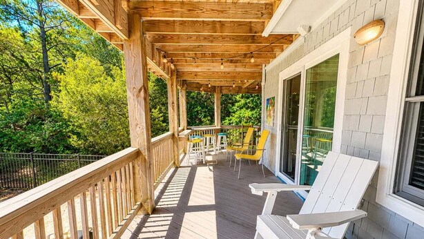 Um deck de madeira com cadeiras Adirondack brancas, uma mesa e um corrimão.