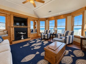 A spacious living room with a fireplace, comfortable seating, large windows with ocean views, and a ceiling fan. - Seven Seas - 4 BR Oceanfront Home w/Private Pool & Ocean Views (Southern Shores)