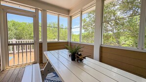 Eine überdachte Terrasse mit einer Holzbank und einem Tisch, großen Fenstern sowie Blick auf eine Außenterrasse und Bäume.