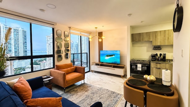 Una sala de estar moderna con sofá, sillón, mesa de comedor y área de cocina, con televisor y electrodomésticos.