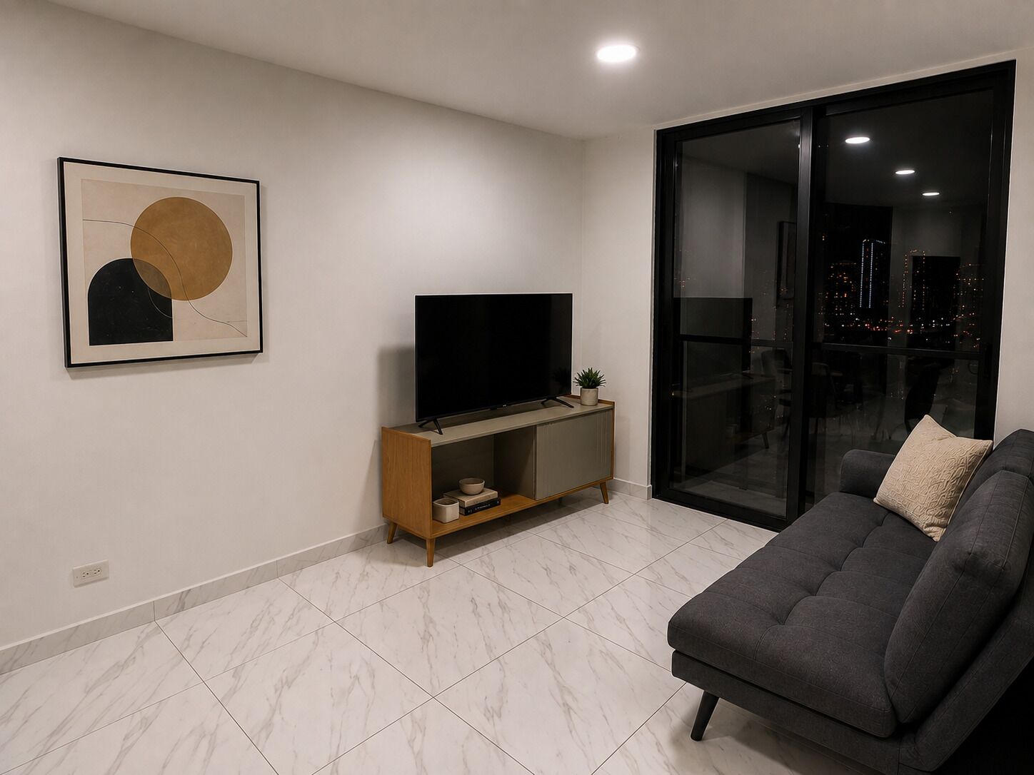 A modern living room with a flat-screen TV on a wooden stand, a grey sofa, and a glass door leading to a balcony with a city view.