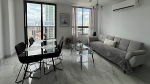 Una sala moderna con una mesa de comedor de vidrio, sillas negras, un sofá gris y un ventanal con vistas a la ciudad.