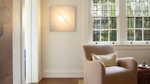 Una habitación con un sillón color crema, una lámpara fijada a la pared y una ventana con vistas a follaje.